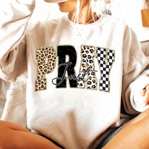 Könnte beinhalten: Cremefarbenes Sweatshirt mit den Worten "PRAY Just" in einer Mischung aus Leopardenmuster, Schwarz und Schachbrettmuster. Das Wort "Just" ist kursiv geschrieben. Ein lässiger, bequemer Kleidungsstil.