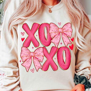 May include: Cream-colored sweatshirt featuring the letters "XOXO" in a bold, pink font with a gold outline. The design includes pink bows with gold trim and small red hearts, creating a Valentine's Day theme.