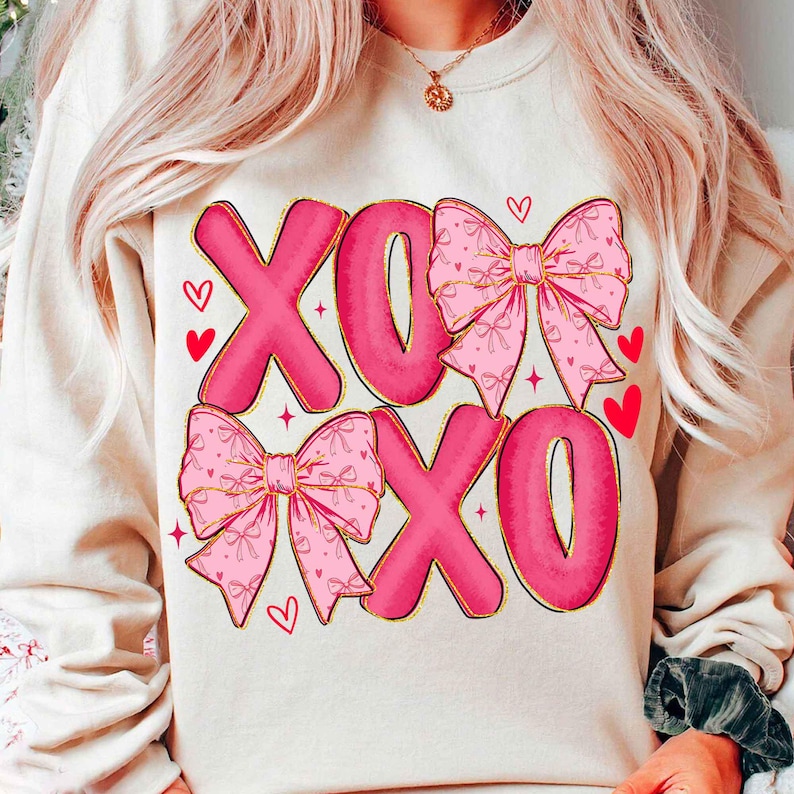 May include: Cream-colored sweatshirt featuring the letters "XOXO" in a bold, pink font with a gold outline. The design includes pink bows with gold trim and small red hearts, creating a Valentine's Day theme.