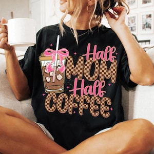 K&ouml;nnte beinhalten: Schwarzes T-Shirt mit braunem und wei&szlig;em Karomuster und dem Text "Half Mom Half Coffee" in Rosa und Braun. Ein rosafarbenes Band befindet sich auf einem Glas mit Eiskaffee und einem Strohhalm.
