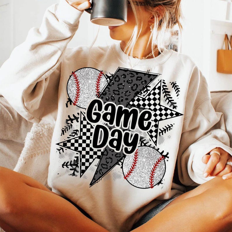 May include: White sweatshirt with a graphic design featuring baseballs, a checkered star, a lightning bolt, and leopard print. The text "Game Day" is printed in black.