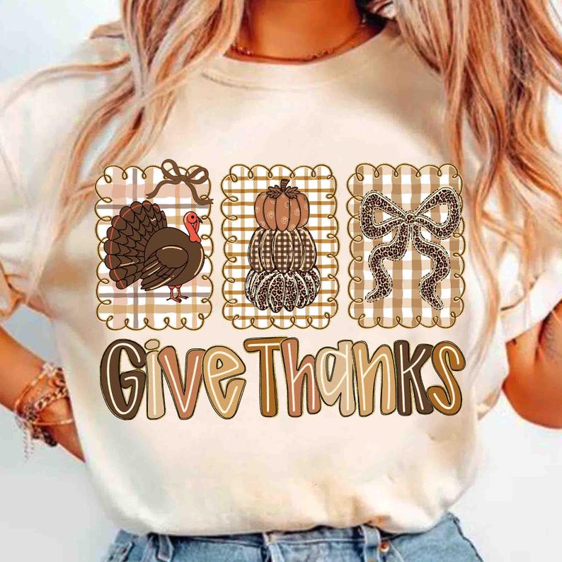 May include: Cream-colored t-shirt featuring three Thanksgiving-themed designs. The designs include a turkey, stacked pumpkins, and a bow, all set against a plaid background. The words "Give Thanks" are printed below the designs.