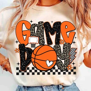 Peut inclure: T-shirt cr&egrave;me avec un motif sur le th&egrave;me du basket-ball. Le design pr&eacute;sente les mots "Game Day Love" en orange, imprim&eacute; l&eacute;opard et noir, ainsi qu'un ballon de basket, un panier et des accents floraux. Un motif &agrave; carreaux noir et blanc est en bas.