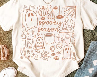 Op de afbeelding: Crèmekleurig T-shirt met een bruine Halloween-afbeelding. Het ontwerp bevat een spook, pompoen, spinnenweb, heksenhoed en de woorden "spooky season" en "if you dare". Een zilveren ketting is zichtbaar.