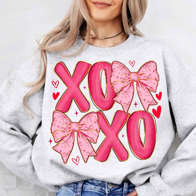 May include: A light gray sweatshirt with the letters "XOXO" in pink, accented with pink bows and red hearts. The bows have gold trim and a delicate pattern. A cute and romantic design.
