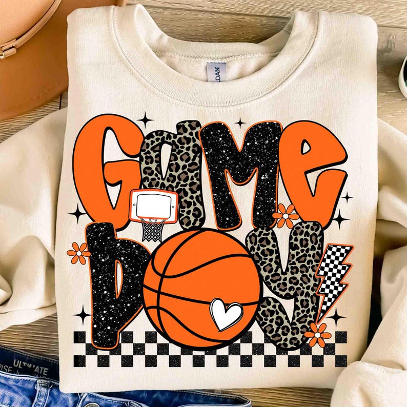 Peut inclure: Sweat-shirt cr&egrave;me avec l'inscription "Game Day Love" en orange, noir et imprim&eacute; l&eacute;opard. Un ballon de basket-ball avec un c&oelig;ur, un panier et un motif &agrave; carreaux compl&egrave;tent le design. Le sweat-shirt est de style d&eacute;contract&eacute; et sportif.