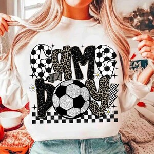 May include: White sweatshirt with a soccer-themed graphic. The design features the words "GAME DAY" in a mix of fonts and patterns, including soccer ball and leopard print. A soccer ball is in the center, with small daisy accents and a checkered border.