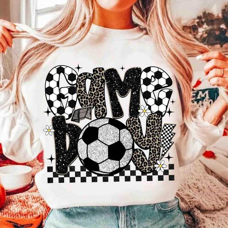 May include: White sweatshirt with a soccer-themed graphic. The design features the words "GAME DAY" in a mix of fonts and patterns, including soccer ball and leopard print. A soccer ball is in the center, with small daisy accents and a checkered border.