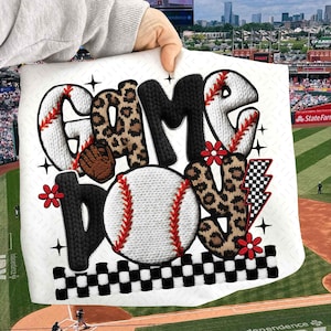 May include: White baseball-themed design with the words "GAME DAY" in a mix of textures and patterns. The letters are in a combination of knitted, leopard print, and baseball stitch designs. Includes a baseball and a baseball glove.