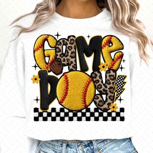 May include: White sweatshirt with the words "GAME DAY" in a playful design. The letters are a mix of yellow, leopard print, and black, with a softball graphic. Accents include flowers, a lightning bolt, and a checkered border.