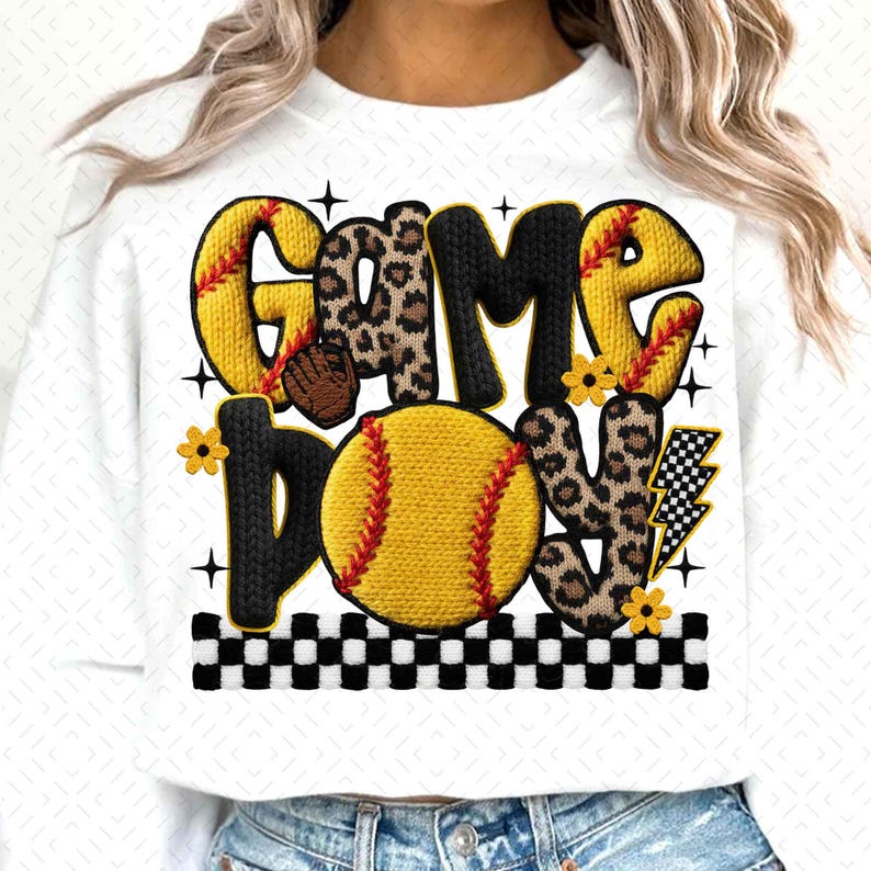 May include: White sweatshirt with the words "GAME DAY" in a playful design. The letters are a mix of yellow, leopard print, and black, with a softball graphic. Accents include flowers, a lightning bolt, and a checkered border.