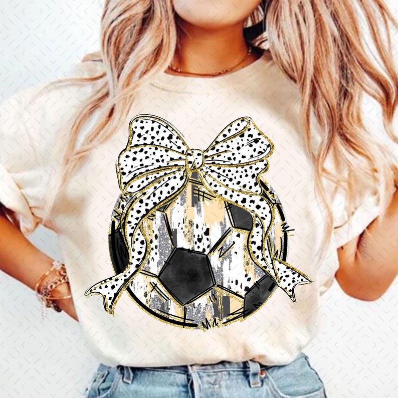 Op de afbeelding: Een wit T-shirt met een zwart-wit voetbalontwerp. De voetbal is versierd met een gouden en zwarte strik.
