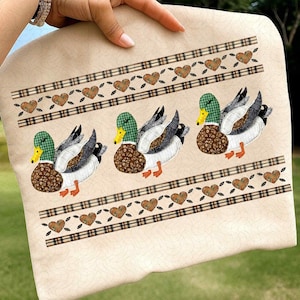 May include: A beige fabric item featuring three duck appliqués with green and brown patterned bodies, white and gray wings, and yellow beaks. The design includes plaid and heart-shaped accents.