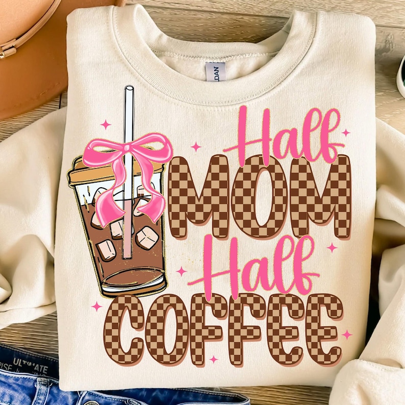 K&ouml;nnte beinhalten: Ein beiger Pullover mit einem braunen und wei&szlig;en Karomuster und dem Text "Half Mom Half Coffee" in rosa Buchstaben. Links neben dem Design befindet sich eine Comic-Abbildung einer Kaffeetasse mit rosa Strohhalm und Marshmallows.