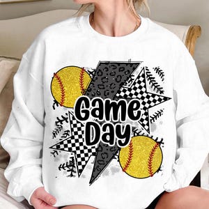 Op de afbeelding: Witte sweatshirt met een "Game Day" afbeelding. Het ontwerp bevat twee gele softballs met rode stiksels, een zwart-wit geblokte ster, een bliksemschicht met luipaardprint en honkbalstikselaccenten.