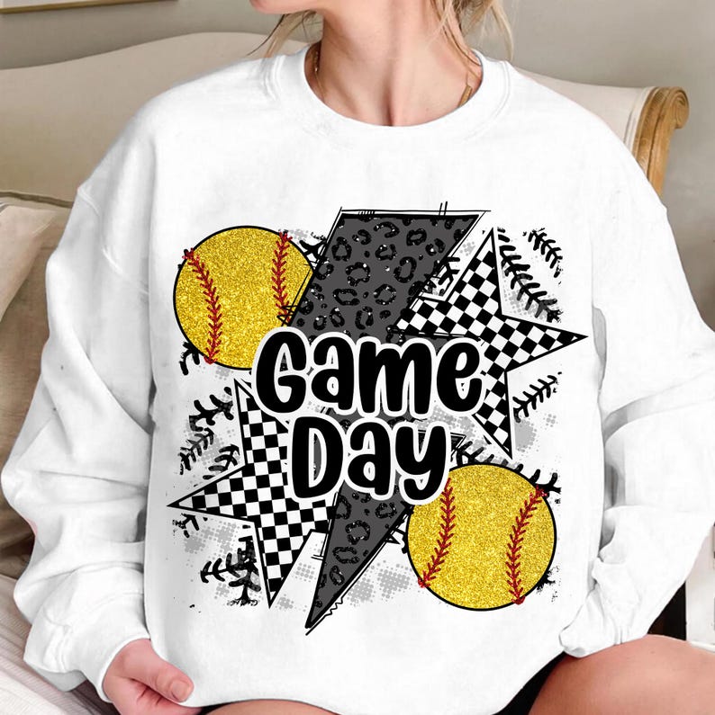 Op de afbeelding: Witte sweatshirt met een "Game Day" afbeelding. Het ontwerp bevat twee gele softballs met rode stiksels, een zwart-wit geblokte ster, een bliksemschicht met luipaardprint en honkbalstikselaccenten.