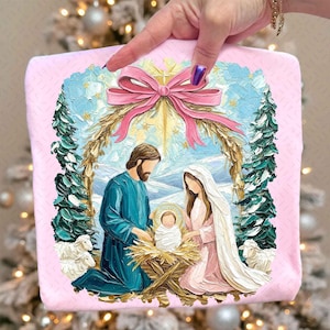 May include: A pink square decorative item featuring a Nativity scene. The artwork depicts Mary, Joseph, and baby Jesus in a manger, surrounded by sheep and evergreen trees. A large pink bow and gold accents frame the scene.