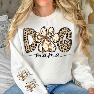 Puede incluir: Sudadera blanca con un dise&ntilde;o de "Dog Mama". La palabra "DOG" y la letra "G" tienen estampado de leopardo. La huella de la pata es marr&oacute;n con un lazo marr&oacute;n. Las mangas tienen huellas de patas y nombres.