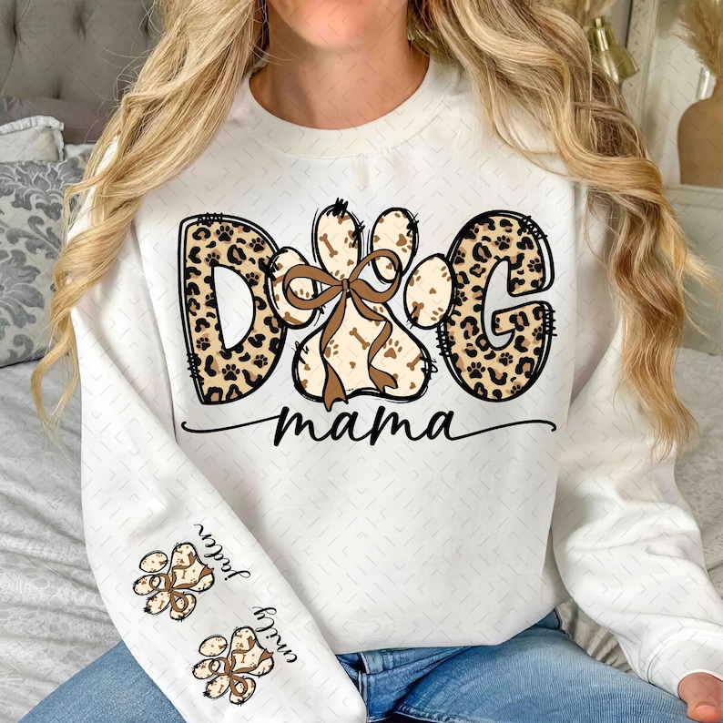 Puede incluir: Sudadera blanca con un dise&ntilde;o de "Dog Mama". La palabra "DOG" y la letra "G" tienen estampado de leopardo. La huella de la pata es marr&oacute;n con un lazo marr&oacute;n. Las mangas tienen huellas de patas y nombres.