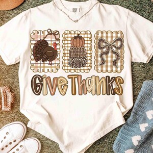 May include: A white t-shirt featuring a Thanksgiving design. The design includes a turkey, stacked pumpkins, and a bow, all within decorative frames. The phrase "Give Thanks" is printed in a stylized font.