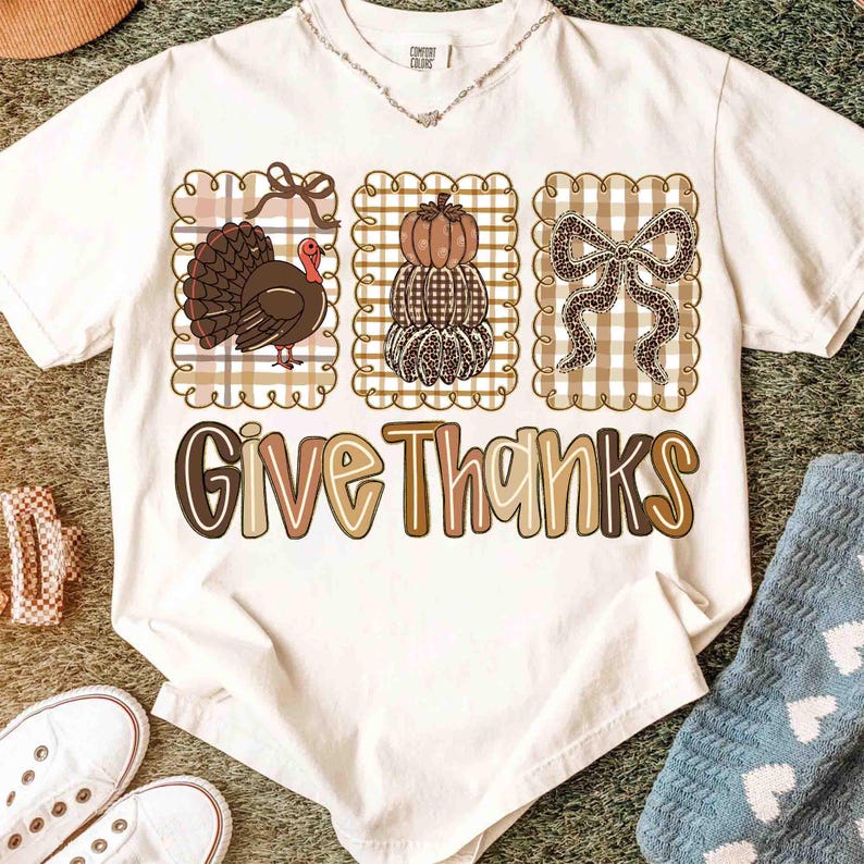 May include: A white t-shirt featuring a Thanksgiving design. The design includes a turkey, stacked pumpkins, and a bow, all within decorative frames. The phrase "Give Thanks" is printed in a stylized font.