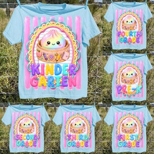 May include: A light blue t-shirt featuring a graphic of a smiling dumpling in a bamboo steamer surrounded by school supplies. The design includes a colourful scalloped frame, pink vertical stripes, and the text Kindergarten in bold letters.