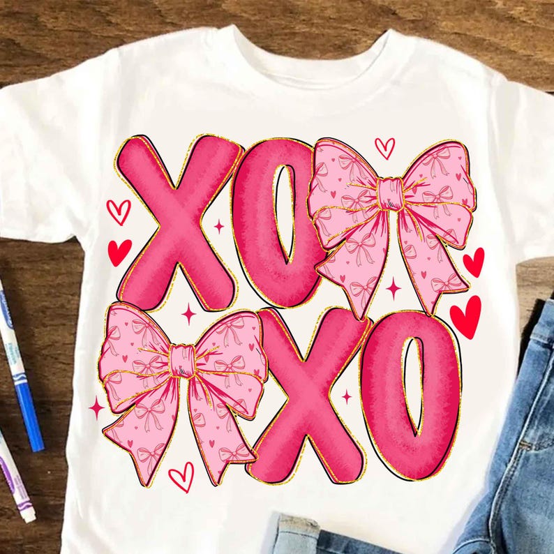 May include: White t-shirt featuring the text "XOXO" in large, pink letters with gold outlines. Pink bows with gold details and red hearts are also featured on the shirt. A cute and festive design.