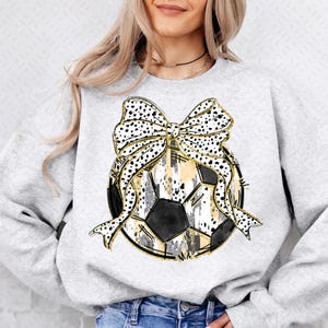Op de afbeelding: Een grijs sweatshirt met een zwart-witte voetbal met een gouden glitterontwerp en een zwart-witte stippenstrik met gouden glitteraccenten.