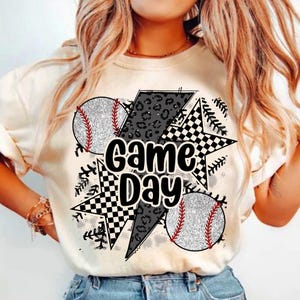 May include: White t-shirt with a graphic design featuring a black and white checkered star, a black lightning bolt, and two baseballs with red stitching. The text "Game Day" is written in black letters.