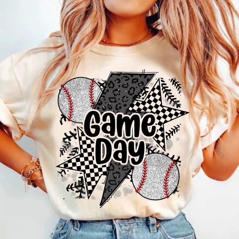 May include: White t-shirt with a graphic design featuring a black and white checkered star, a black lightning bolt, and two baseballs with red stitching. The text "Game Day" is written in black letters.