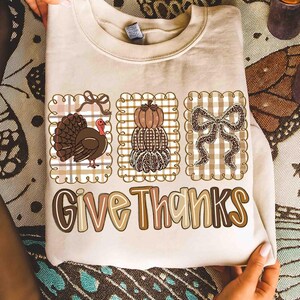 May include: Beige sweatshirt with three decorative squares featuring a turkey, stacked pumpkins, and a bow. The words "Give Thanks" are printed in a fall-themed font. The design is in shades of brown, tan, and cream.