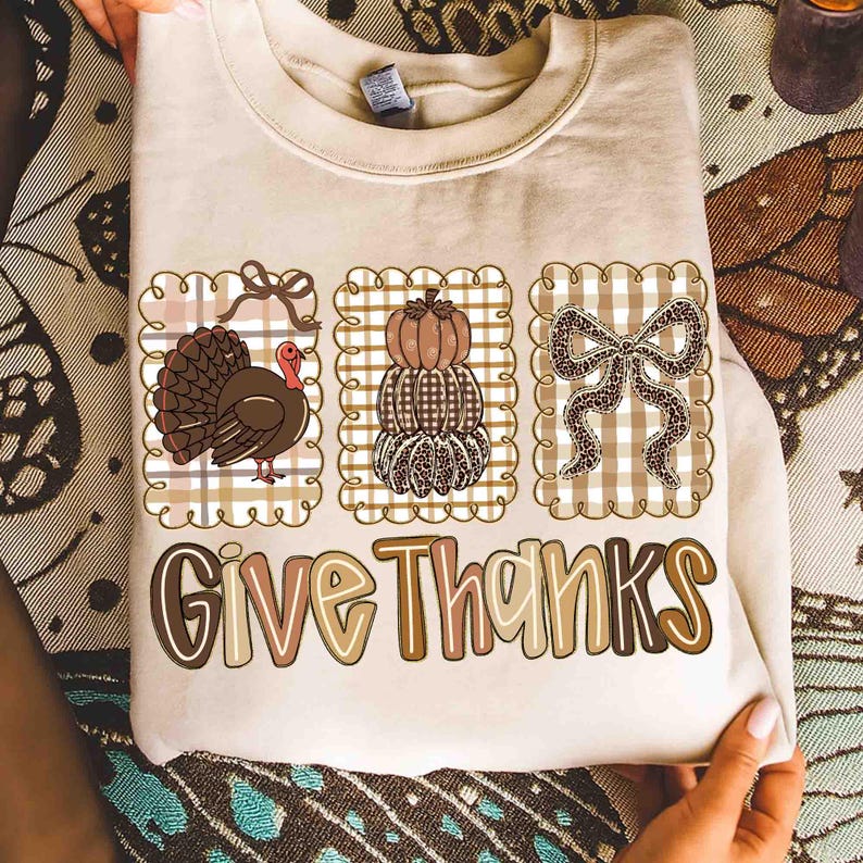May include: Beige sweatshirt with three decorative squares featuring a turkey, stacked pumpkins, and a bow. The words "Give Thanks" are printed in a fall-themed font. The design is in shades of brown, tan, and cream.