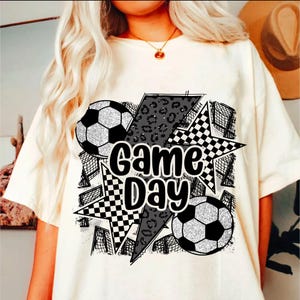 May include: White t-shirt with a black and white graphic design featuring the text "Game Day" and soccer balls. The graphic includes a checkered pattern, a leopard print, and a lightning bolt.