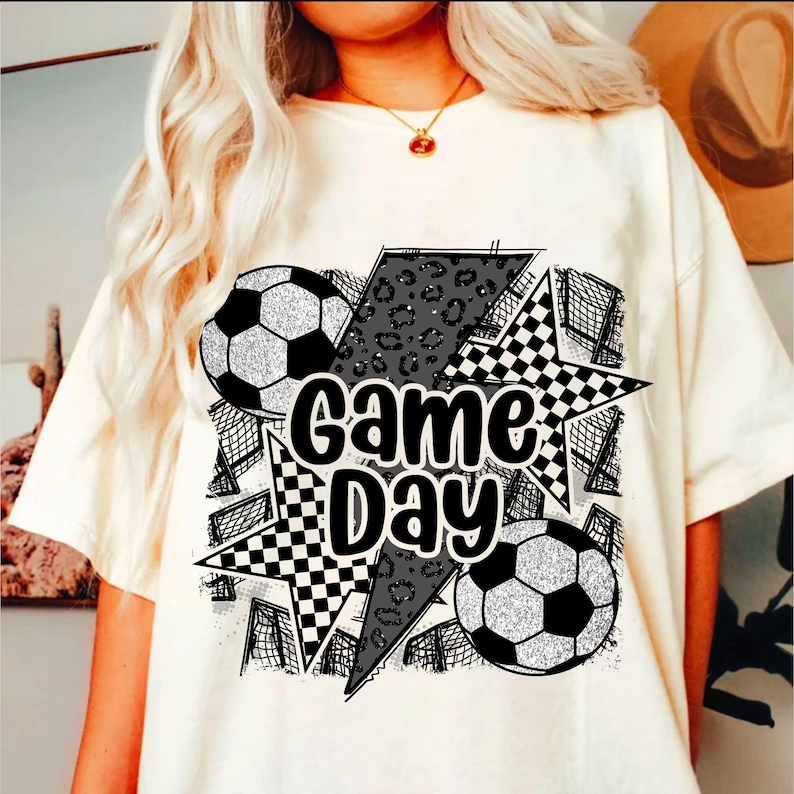 May include: White t-shirt with a black and white graphic design featuring the text "Game Day" and soccer balls. The graphic includes a checkered pattern, a leopard print, and a lightning bolt.