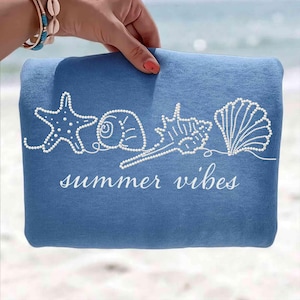 May include: A blue beach towel with white seashell designs and the words "summer vibes" in a script font. The towel features a starfish, a spiral shell, a conch shell, and a scallop shell. The towel is held by a person.