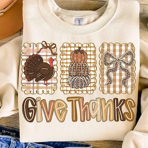 May include: Cream-colored sweatshirt with a Thanksgiving theme. Features three framed images: a turkey, stacked pumpkins, and a bow, all on a plaid background. The words "Give Thanks" are printed in a decorative font.