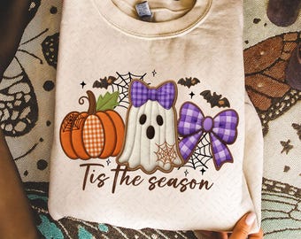May include: Cream-colored sweatshirt featuring a Halloween-themed design. The design includes a pumpkin, a ghost with a purple plaid bow, and a large purple plaid bow. The text "Tis the season" is printed below the graphics.