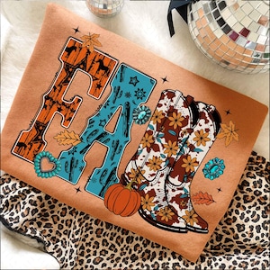 May include: A tan sweatshirt with the word "FALL" in a western-style font. The "F" is orange with horse silhouettes, the "A" is turquoise with cactus designs, and the "LL" is brown with floral cowboy boots. A pumpkin and turquoise accents complete the design.