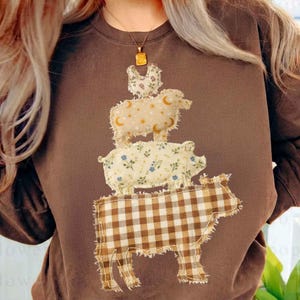 May include: Brown sweatshirt featuring a stacked farm animal design. The design includes a cow, pig, sheep, and chicken, each with unique patterned fabrics. The cow is a brown and white checkered pattern, the pig has a floral pattern, and the sheep has a star and moon pattern.