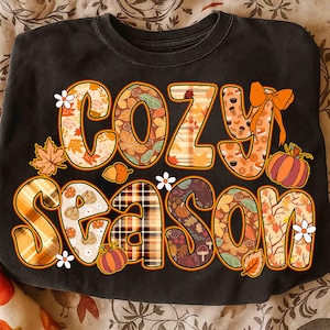 Puede incluir: Sudadera negra con la frase "COZY SEASON" en un diseño otoñal. Las letras están rellenas de patrones de hojas, calabazas y cuadros. Flores blancas y un lazo naranja acentúan el diseño.