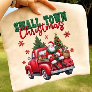 May include: A beige sweatshirt featuring a festive Christmas design. The design includes a red truck carrying gifts, Santa Claus, a Christmas tree, and the text "SMALL TOWN Christmas" in green and red.