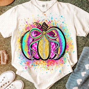 Puede incluir: Camiseta color crema con un diseño de calabaza colorido. La calabaza tiene un centro dorado, un estampado de leopardo rosa y un degradado turquesa y morado. Un gran lazo multicolor adorna la parte superior, con un efecto de pintura salpicada.