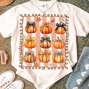May include: White t-shirt featuring a fall-themed design. The design includes nine orange pumpkins with various patterned bows, set against a striped background. A brown border surrounds the pumpkins, with fall leaves and flowers.