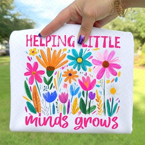May include: White rectangular pillow with a colorful floral design and the text "HELPING LITTLE MINDS GROWS" in pink. The flowers are in various colors, including orange, pink, blue, and yellow.