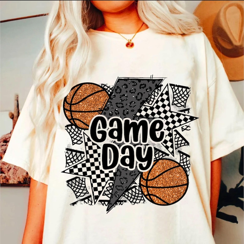 May include: White t-shirt with a graphic design featuring two basketballs, a lightning bolt, and the text "Game Day" in black.