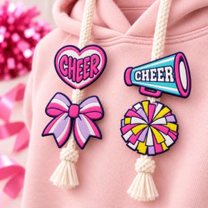 May include: A light pink hooded sweatshirt featuring two thick, cream-colored braided drawstrings. Attached to the drawstrings are four rubber charms: a heart with the word CHEER, a bow, a megaphone with the word CHEER, and a pom-pom.