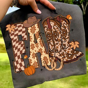 May include: Dark grey t-shirt with the word "FA" in brown and tan autumn designs. The "F" is checkered, and the "A" features oak leaves. A cowboy hat, boots, pumpkin, and horseshoe complete the autumn theme.
