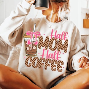 K&ouml;nnte beinhalten: Ein wei&szlig;er Pullover mit einem braunen und wei&szlig;en Karomuster und dem Text "Half Mom Half Coffee" in Pink. Ein rosa Band ist um einen braunen Eiskaffeebecher mit Strohhalm gebunden.