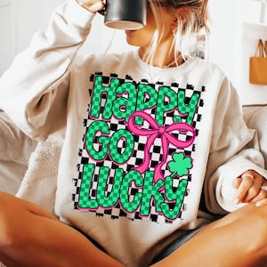May include: White sweatshirt with a checkered pattern in black and green. The text "Happy Go Lucky" is written in green letters with a pink bow and a green shamrock.