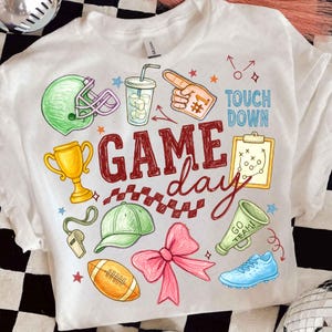 Puede incluir: Camiseta blanca con un diseño colorido de "Game Day". El diseño incluye un casco de fútbol americano, un trofeo, un balón de fútbol y otros gráficos deportivos. También aparecen las palabras "Touch Down" y "Go Team".